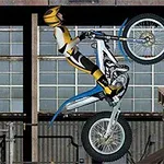 Egy motoros sárga-fekete védőruhában egykeréken hajt egy fehér és kék motorbiciklit egy raktár hátterében